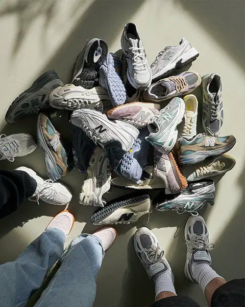 A pile of various New Balance sneakers on a floor, with two pairs of feet in sneakers framing the scene.
