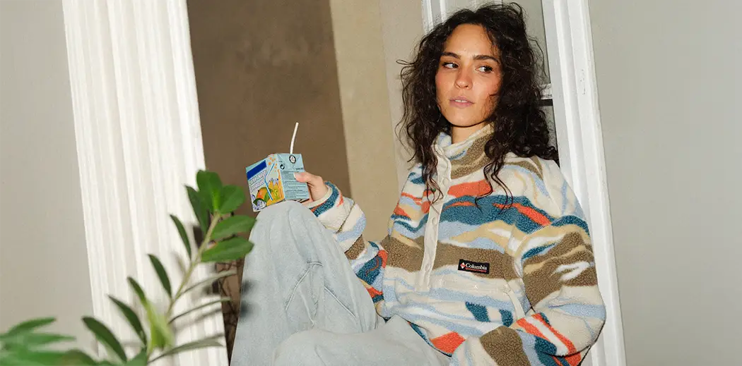 Woman in a colorful fleece jacket sits casually on a windowsill, holding a juice box. She looks thoughtfully outside. Indoor plants in foreground. Cozy mood.