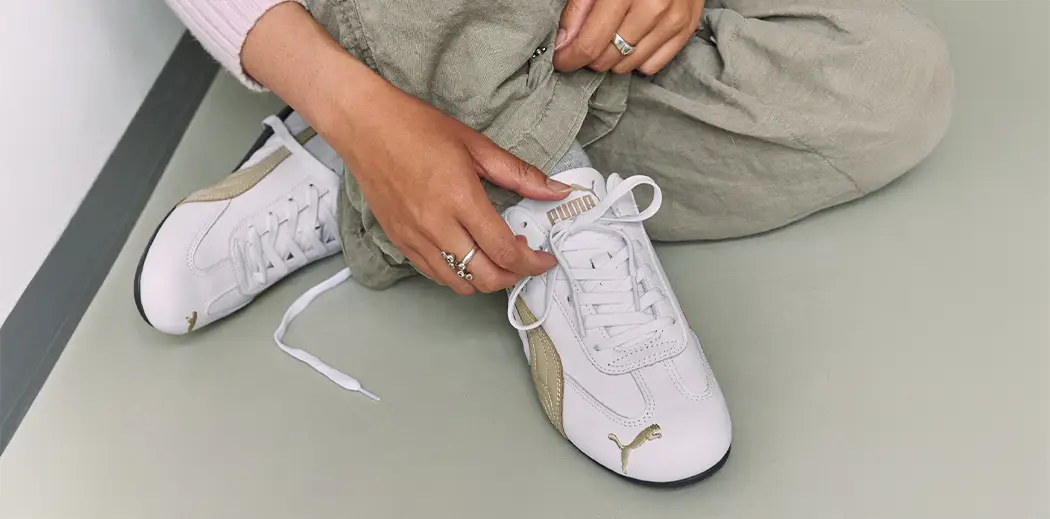 A person wearing a pink sweater and gray pants sits on the floor adjusting their white Puma Speedcat sneakers. The scene conveys a casual, relaxed vibe.